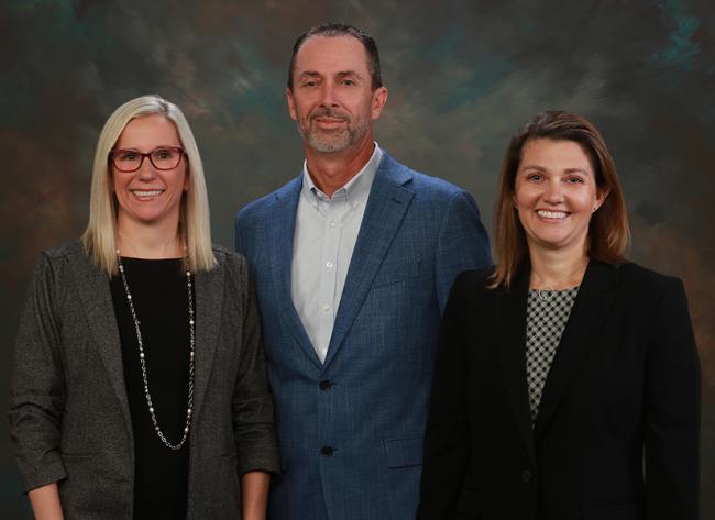 Three business professionals stand together in front of a colorful painting, engaged in conversation.